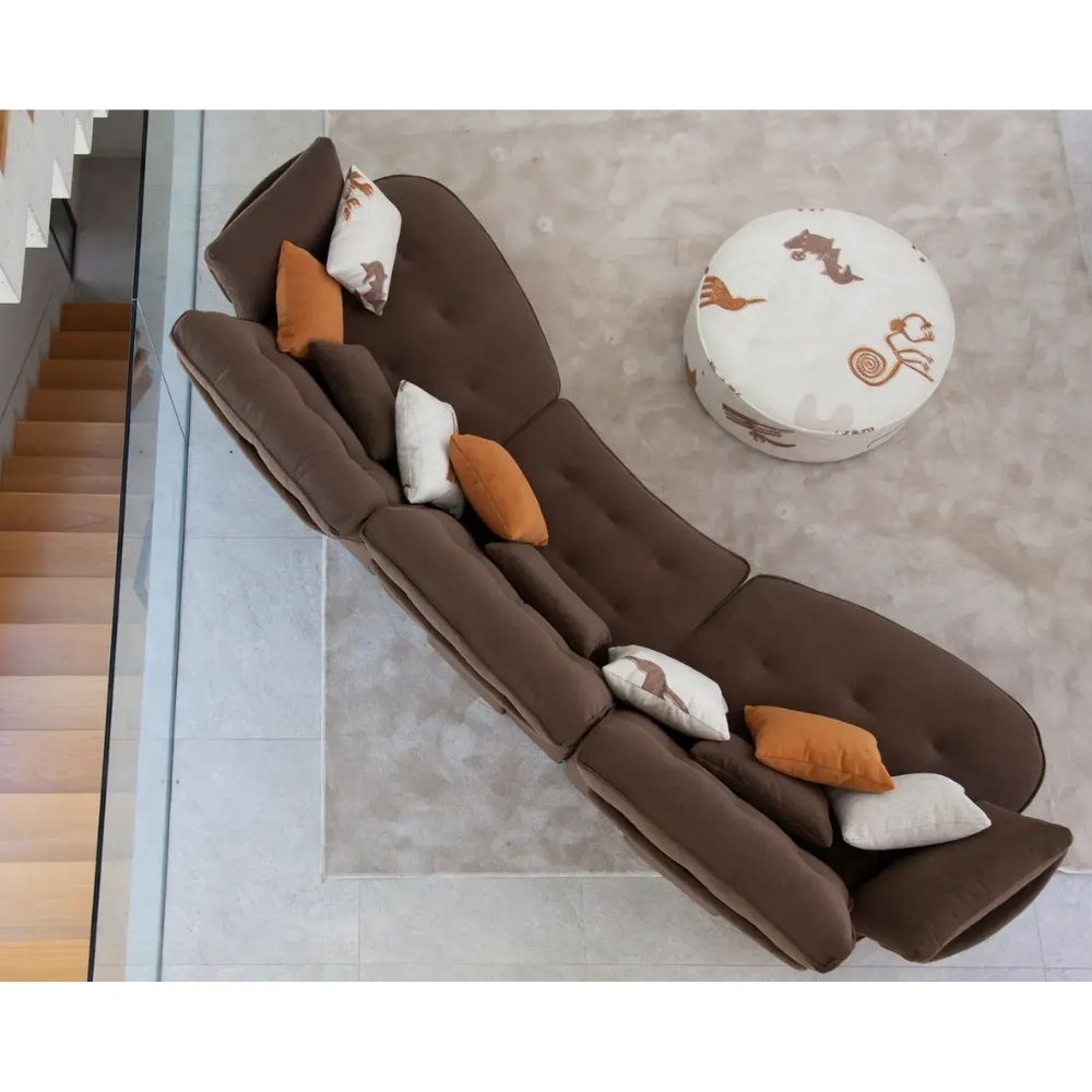 A brown tufted chaise lounge chair with accent pillows, shown in a modern interior setting with a staircase and a round ottoman.