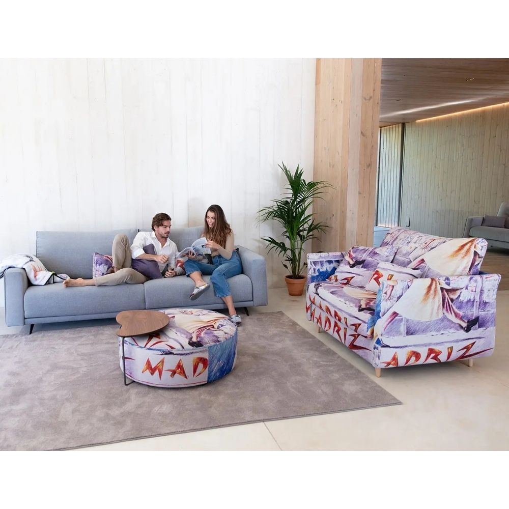 A modern living room scene showing a light blue sofa with two people sitting on it, a matching round ottoman with colorful printed cushions, and a separate upholstered armchair with a bold purple, white, and red graphic print. The room has light wood paneling, a gray rug, and a potted plant.