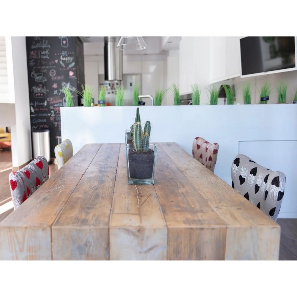 A rustic wooden dining table in a bright modern kitchen setting, styled with a small potted cactus in the center and patterned chairs with red and black heart designs.
