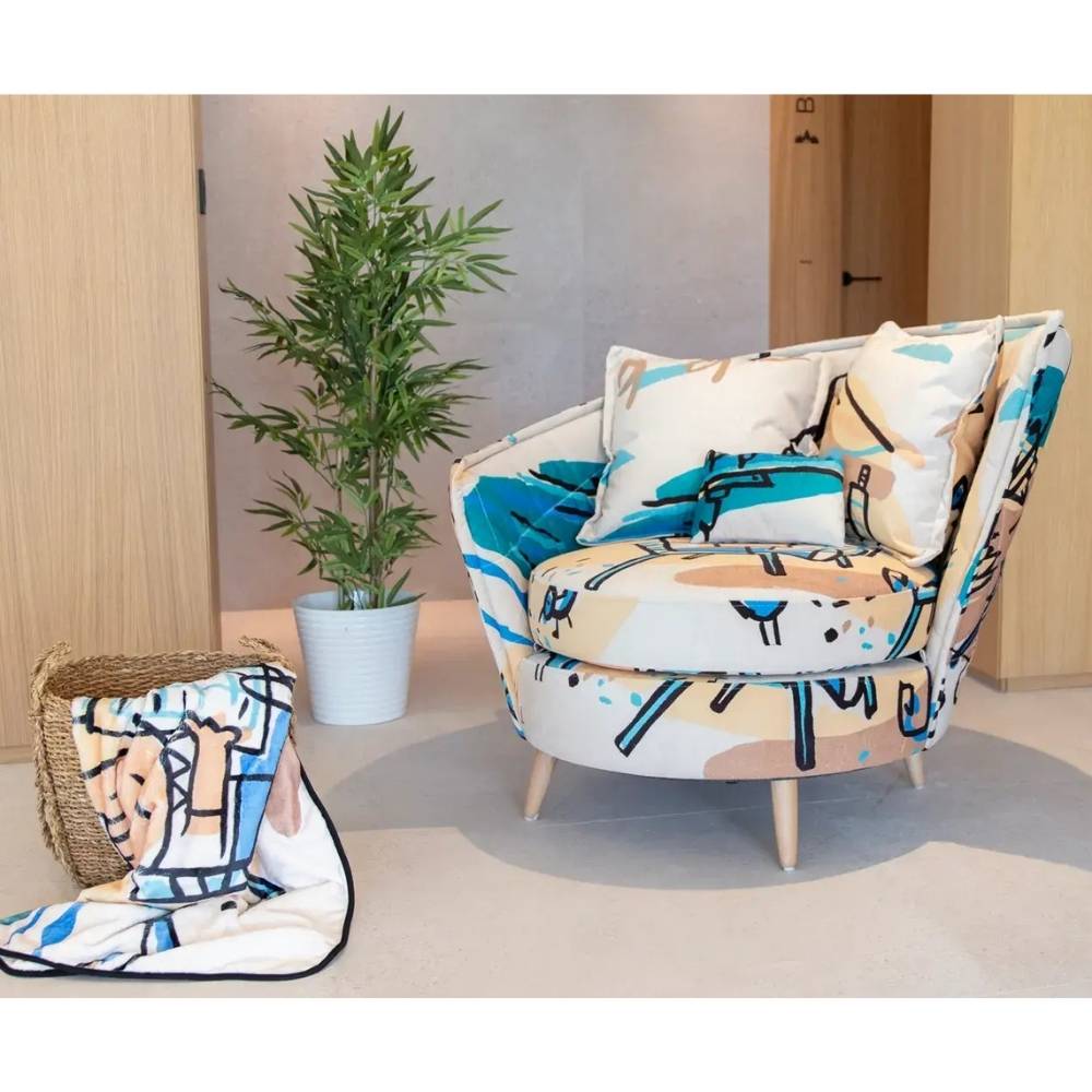 A modern upholstered armchair with a colorful abstract pattern in blue, beige, and white, styled in a room setting next to a potted plant and a woven basket holding a matching throw blanket.