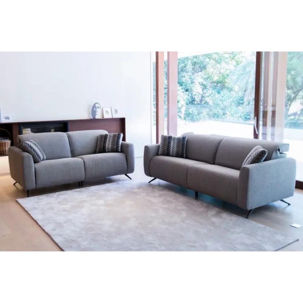 A modern living room scene showing two gray upholstered sofas placed on a large light rug near tall windows. The set includes a two-seat sofa on the left and a three-seat sofa on the right, both with dark legs and patterned throw pillows.