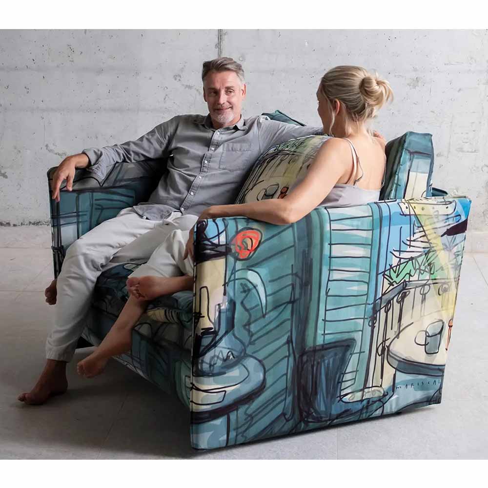 Two people sitting on a colorful, artfully designed sofa against a gray wall.