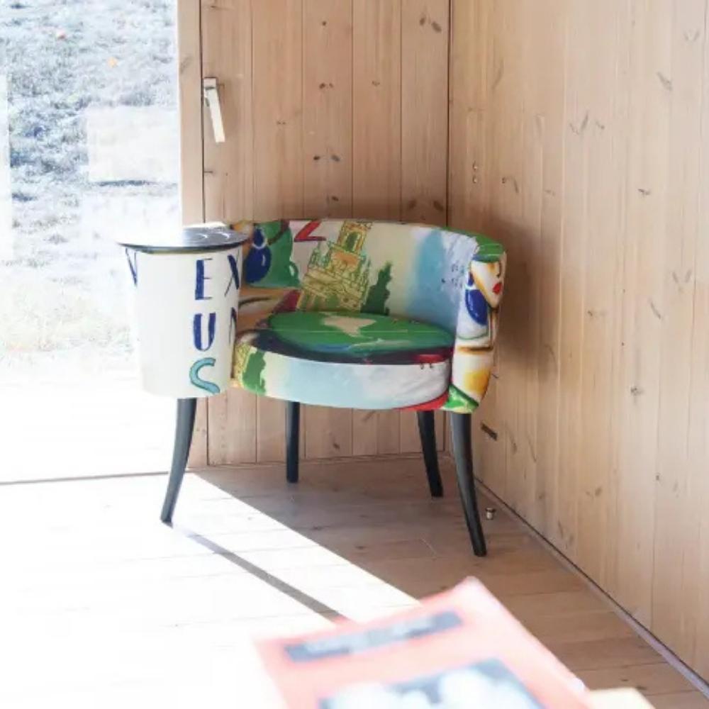 A small upholstered armchair with a colorful abstract and scenic print, with a built in matching cylindrical side table, set against a light wood-paneled wall in a sunlit room.