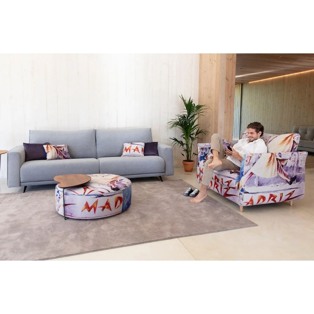 A man relaxes in a modern armchair upholstered in a colorful graffiti-style print featuring the word 'MADRID' and red graffiti lettering. The scene is set in a bright, contemporary living room with a matching round ottoman and a light gray sofa in the background.