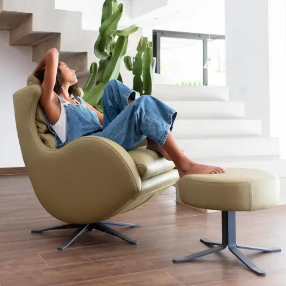 A woman relaxes reclining in a modern beige leather swivel lounge chair with an attached matching footstool, set in a bright contemporary room with white stairs and a cactus.