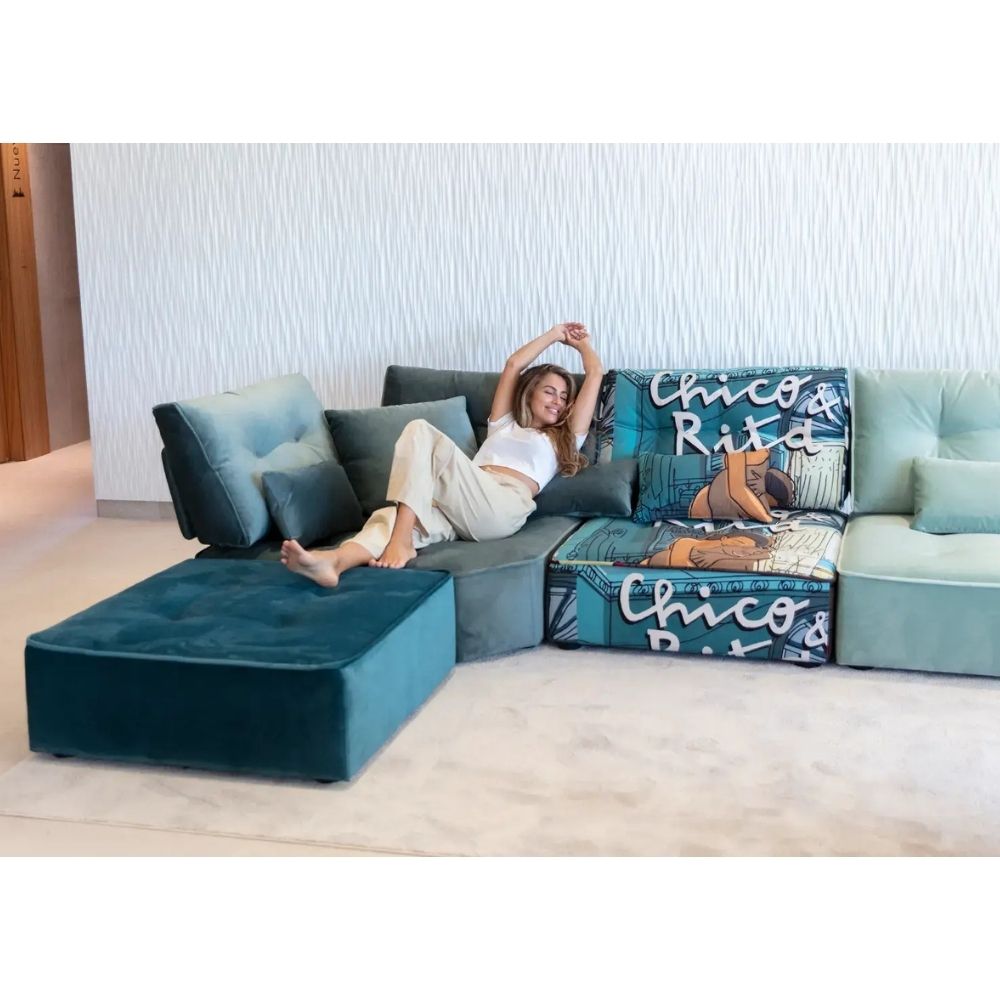 A woman reclines on a modern sectional sofa in a bright indoor setting. The sofa features a dark teal velvet chaise lounge, matching accent pillows, and a patterned seat cushion with blue, teal, and white graphic text and imagery.