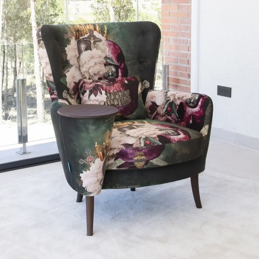 A wingback armchair with a dark green and purple gothic floral print, featuring a matching side table attached to the left arm, set in a modern room with a glass window and brick wall.