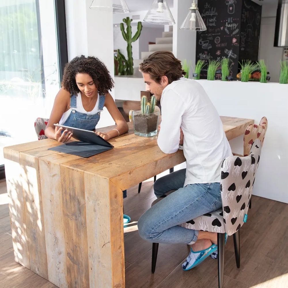 A man and a woman sit at a rustic wooden table in a bright, modern kitchen, looking at a tablet. The woman wears denim overalls, and the man wears a white long-sleeve shirt and jeans.