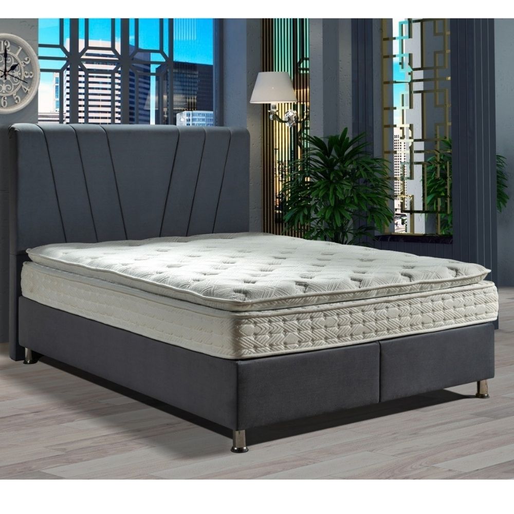 A modern ottoman divan bed frame with a tufted gray upholstered headboard and base, paired with a white tufted mattress, shown in a styled bedroom setting with a window, city view, and plants.