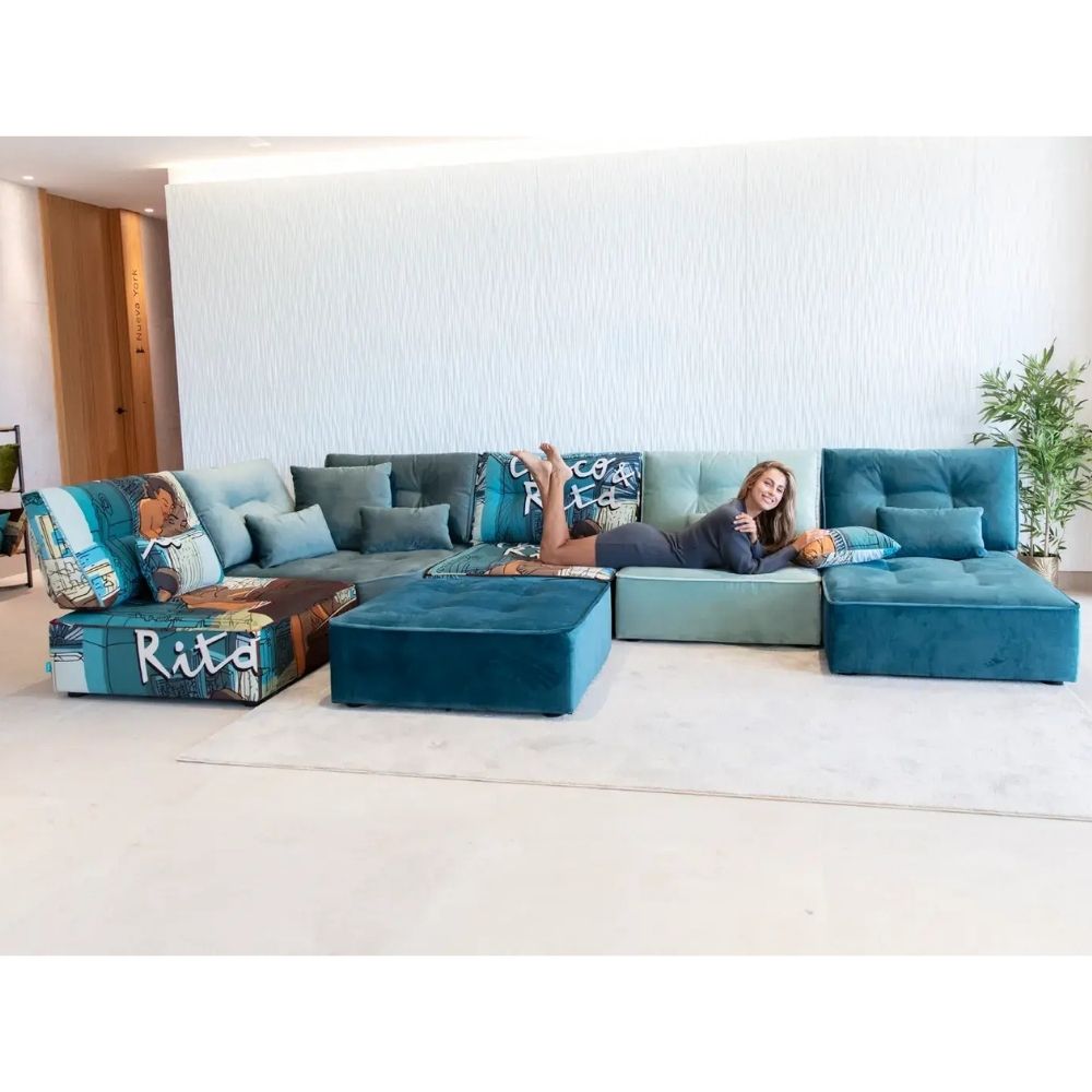 A woman lies on a large sectional sofa in a bright, modern living room. The sectional is upholstered in shades of blue and teal, with matching footstool and decorative throw pillows featuring graphic prints.