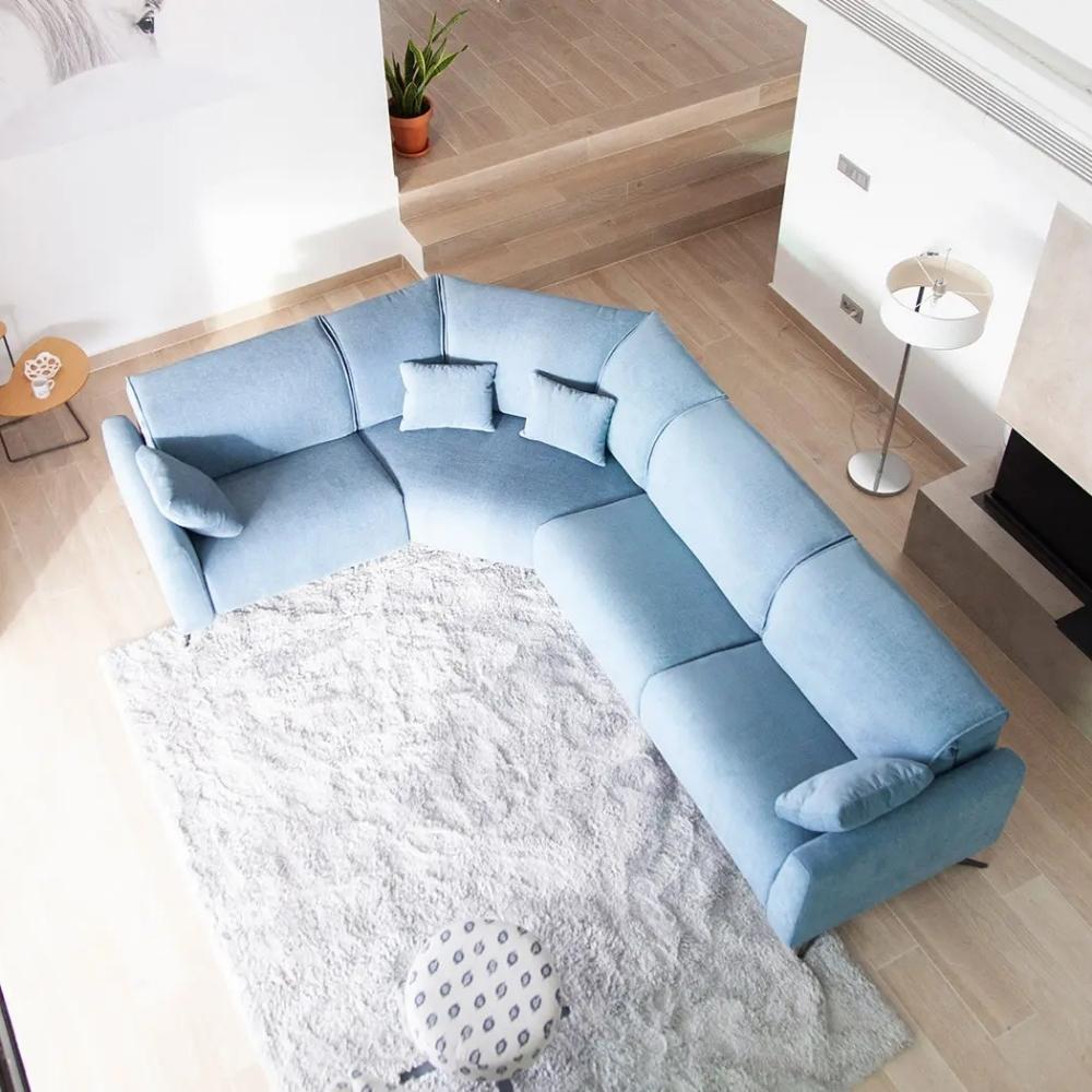 A light blue modular J-shaped sectional sofa with matching throw pillows, styled on a white shag rug in a modern living room with light wood floors.