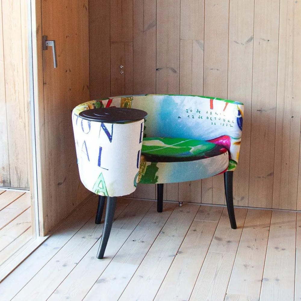 A modern curved lounge chair with a colorful abstract painted fabric and a matching cylindrical side table, set in a room with light wood paneled walls and floor.