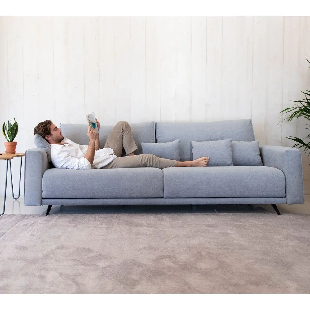 A man relaxes on a light blue fabric sofa, reading a book. The scene is set in a modern living room with a white paneled wall, a gray rug, a small side table with a potted plant, and a green plant in the background.