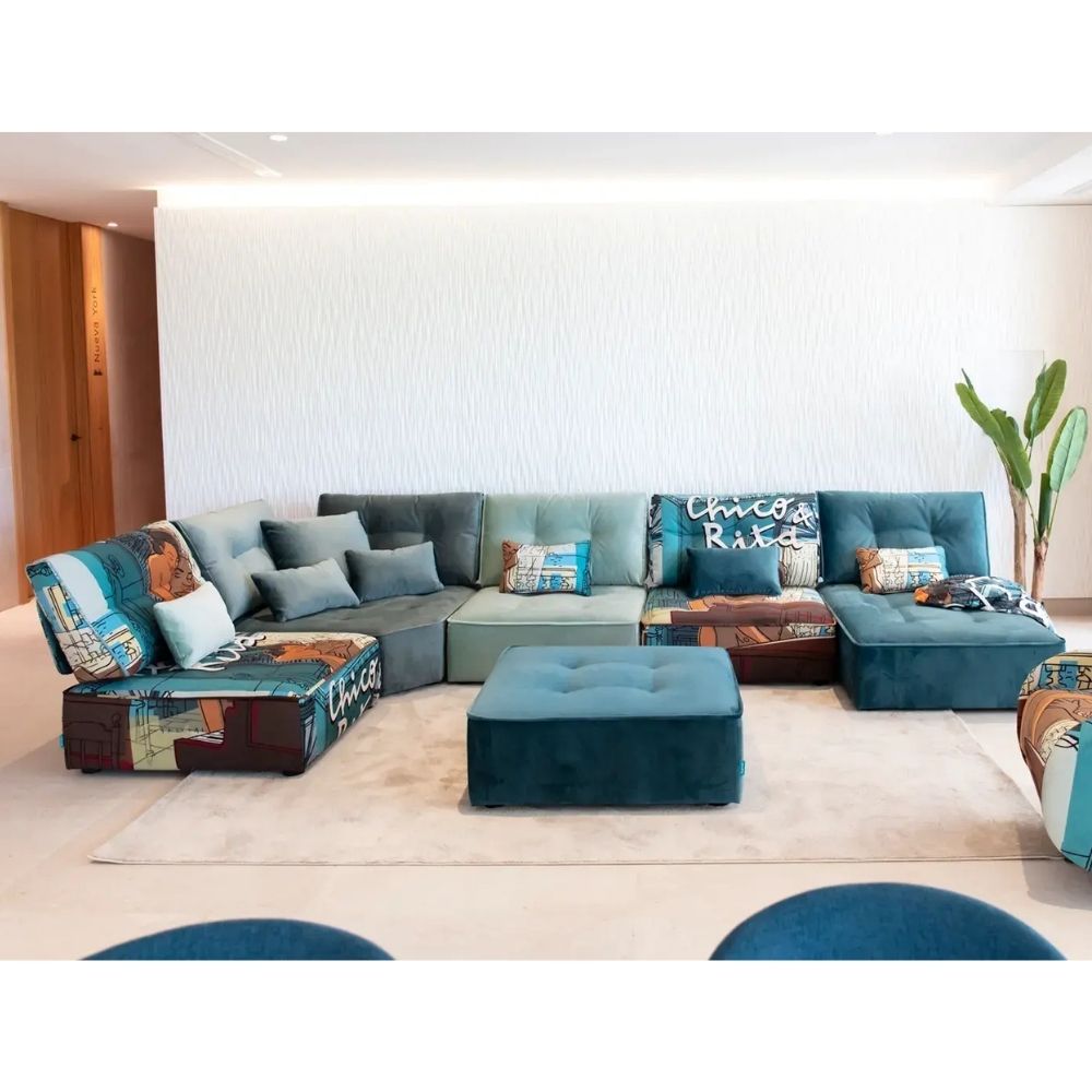 A modern sectional sofa set in shades of teal, blue, and gray, styled with patterned throw pillows and a matching tufted footstool in a bright, contemporary living room.