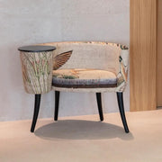 A modern accent chair with a curved backrest, upholstered in a beige and brown botanical patterned fabric, featuring black tapered legs and a matching cylindrical side table on black legs.