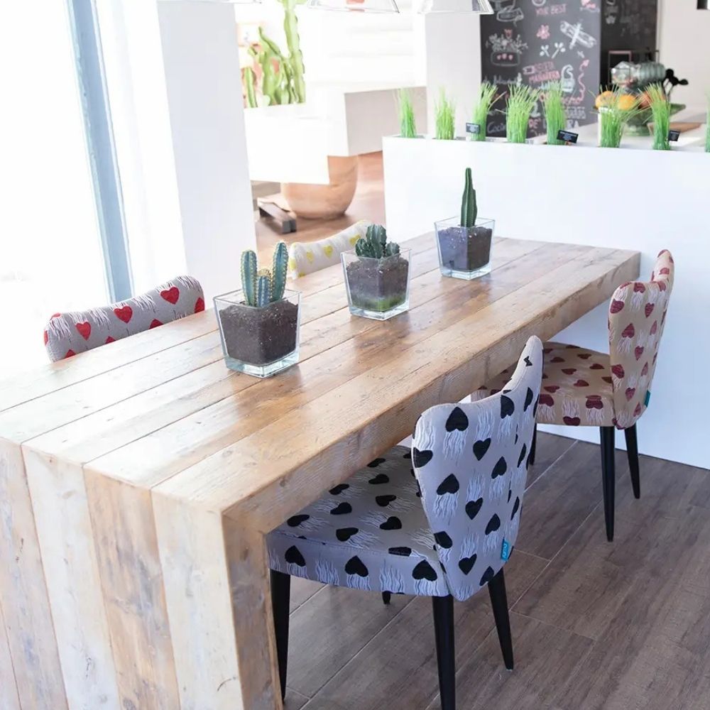A rustic light wood dining table in a bright modern room, surrounded by patterned chairs including one with black hearts on white and another with red hearts on beige. Three small potted cacti sit on the table.