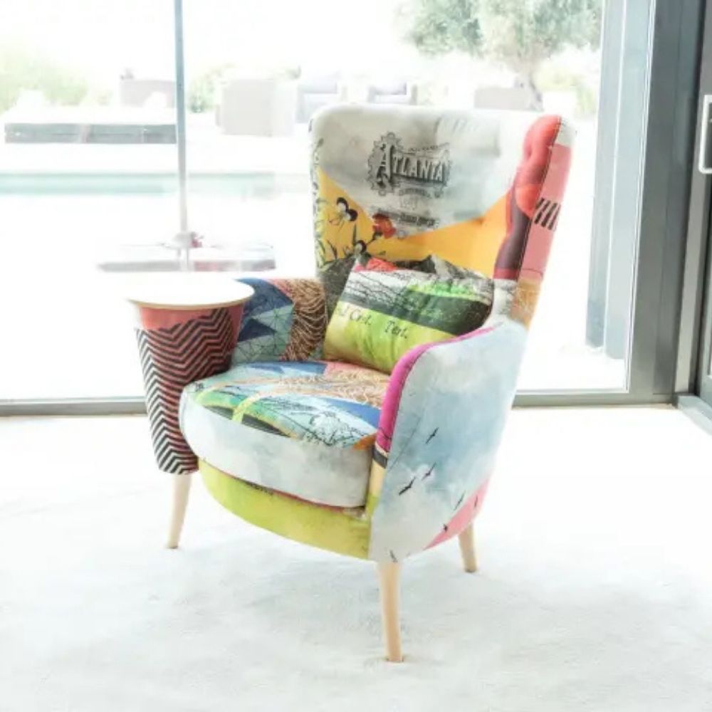 A colorful patchwork wingback armchair with a side table built into the arm, featuring a collage of vintage travel illustrations and patterns, set in a bright modern room with a glass door.