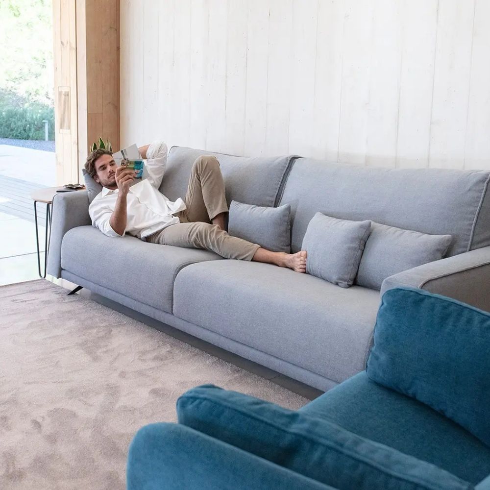 A man relaxes on a light gray modular sofa in a modern room, holding a phone. The scene includes a blue armchair in the foreground, a beige rug, and a light wood-paneled wall.