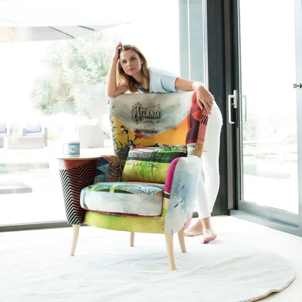 A woman in a light blue top and white pants leans against a highly colorful, patterned armchair with built in side table in one arm. in a bright, modern room with large glass doors.