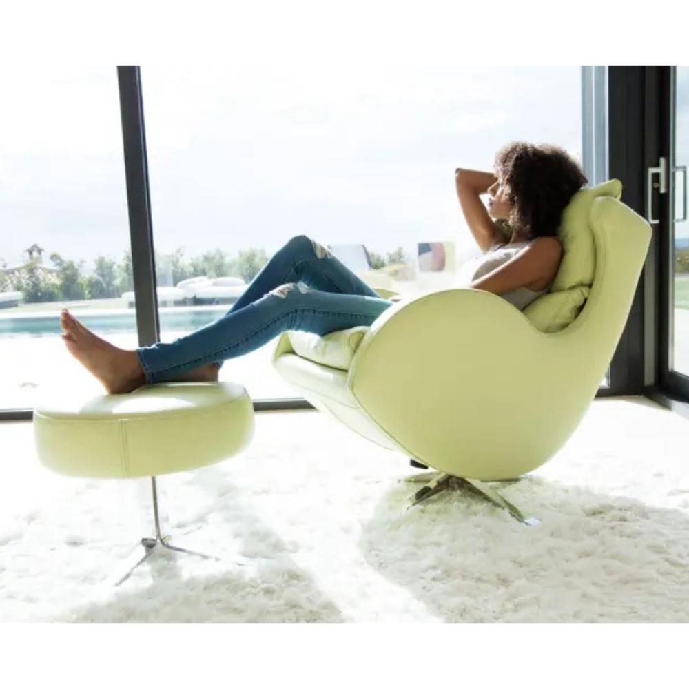 A woman relaxes in a modern pale yellow-green lounge chair with her feet resting on a matching footstool, set in a bright room with large windows and a white shag rug.