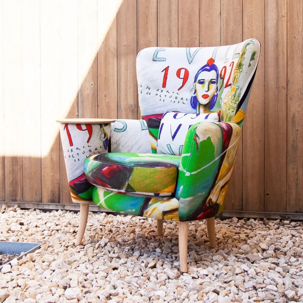 A high-back upholstered armchair with colorful abstract and graphic artwork printed across the seat and back, with a side table built into the arm, set on a bed of small white stones in front of a wooden fence.