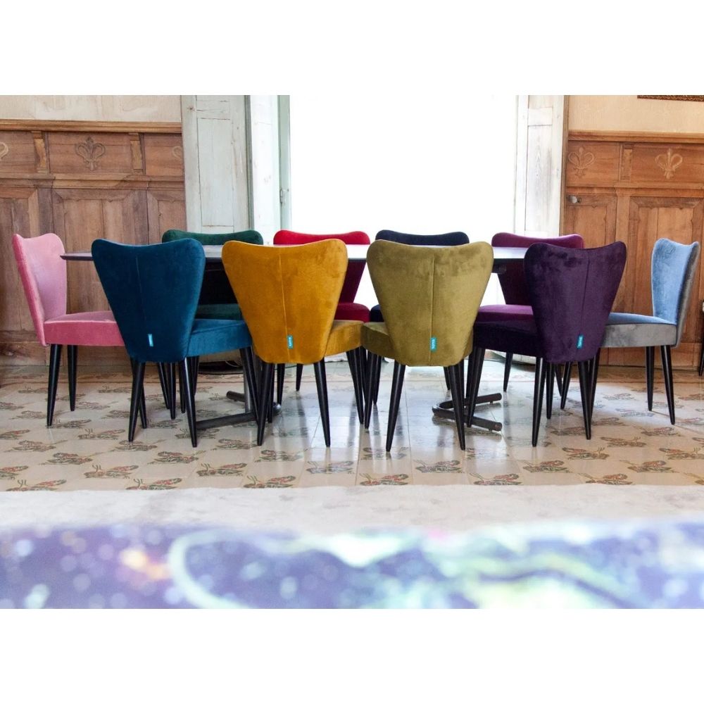 A set of colorful velvet dining chairs arranged around a table in a vintage room with wood-paneled walls and a patterned tile floor.