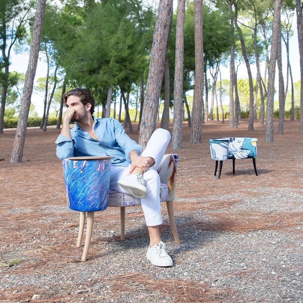 A man sits outdoors in a wooded park setting on a decorative armchair with built in side table, wearing a light blue shirt and white pants. Another matching chair is visible in the background.