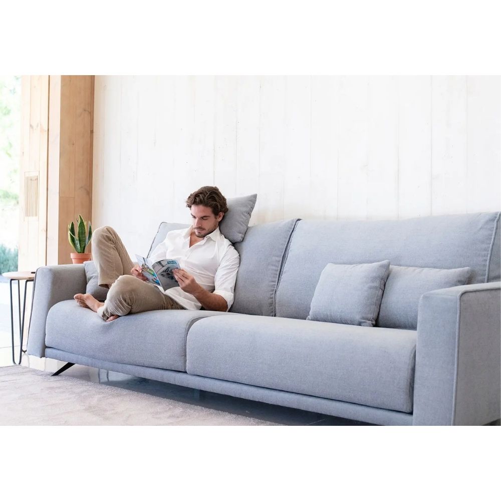 A man relaxes on a light gray fabric sofa in a bright, modern living room, holding a book and laptop. The sofa has three matching throw pillows and is positioned against a white paneled wall.