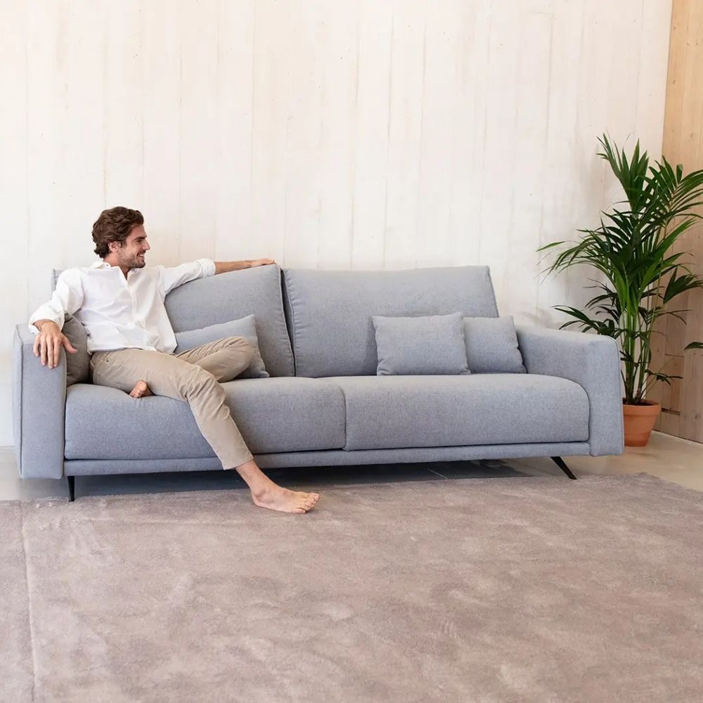 A man in a white shirt and beige trousers sits barefoot on a light gray fabric sofa with matching throw pillows in a bright, modern room with a large gray rug and a potted plant.