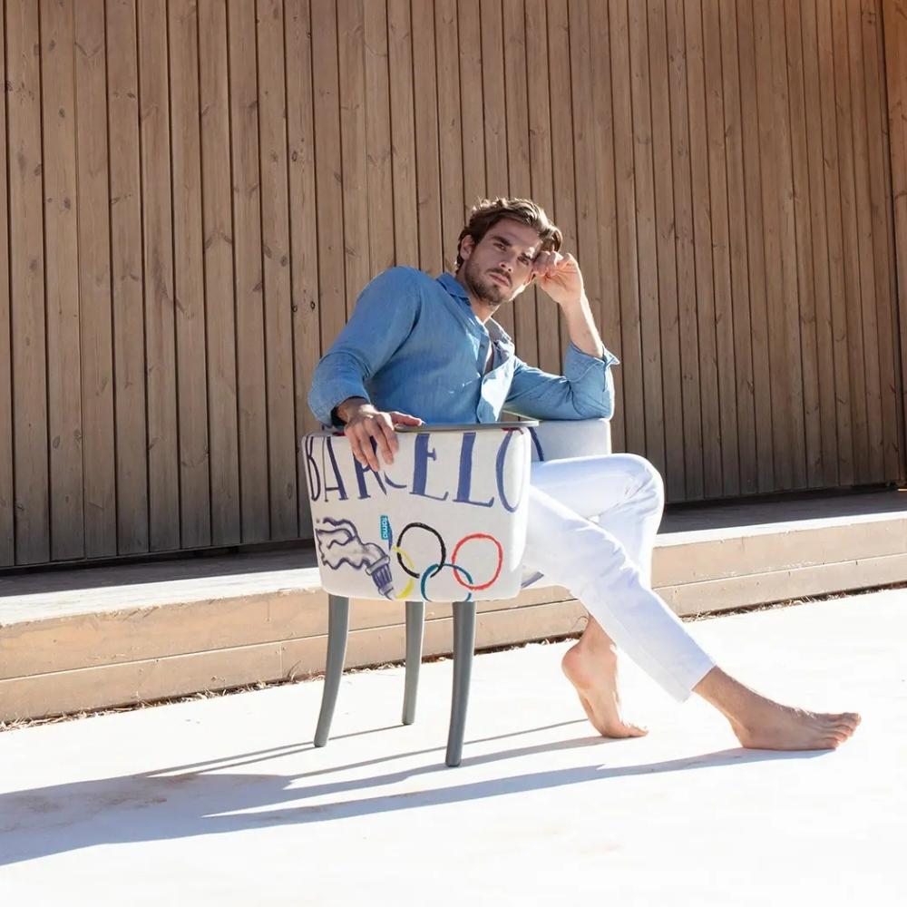 A man in a blue shirt and white pants sits barefoot on an accent chair with 'BARCELONA' and the Olympic rings printed on the fabric, posed in front of a wooden wall.