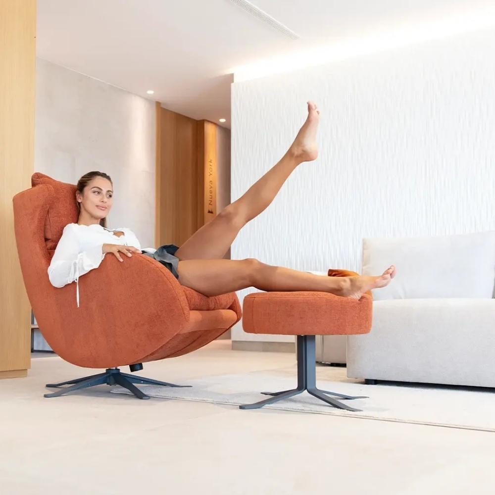 A woman reclines in a modern orange upholstered swivel armchair with a matching footrest in a bright, contemporary living room. The chair and ottoman have dark metal star bases.