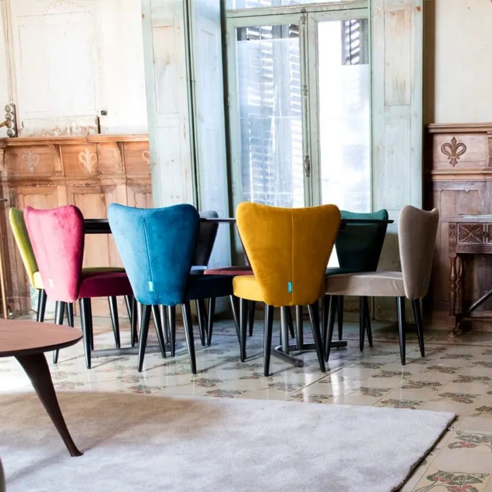 A set of colorful velvet dining chairs in pink, blue, yellow, green, and beige, arranged around a dark table in a vintage-style room with tiled floors and wooden cabinets.