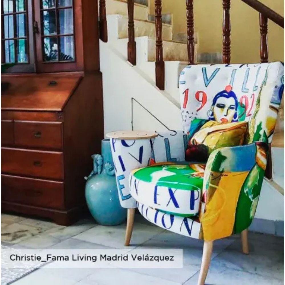A highly decorative, colorful armchair with a graffiti-style art print featuring a woman's face and text, with a built in side table, styled in a home interior next to a wooden cabinet and staircase.