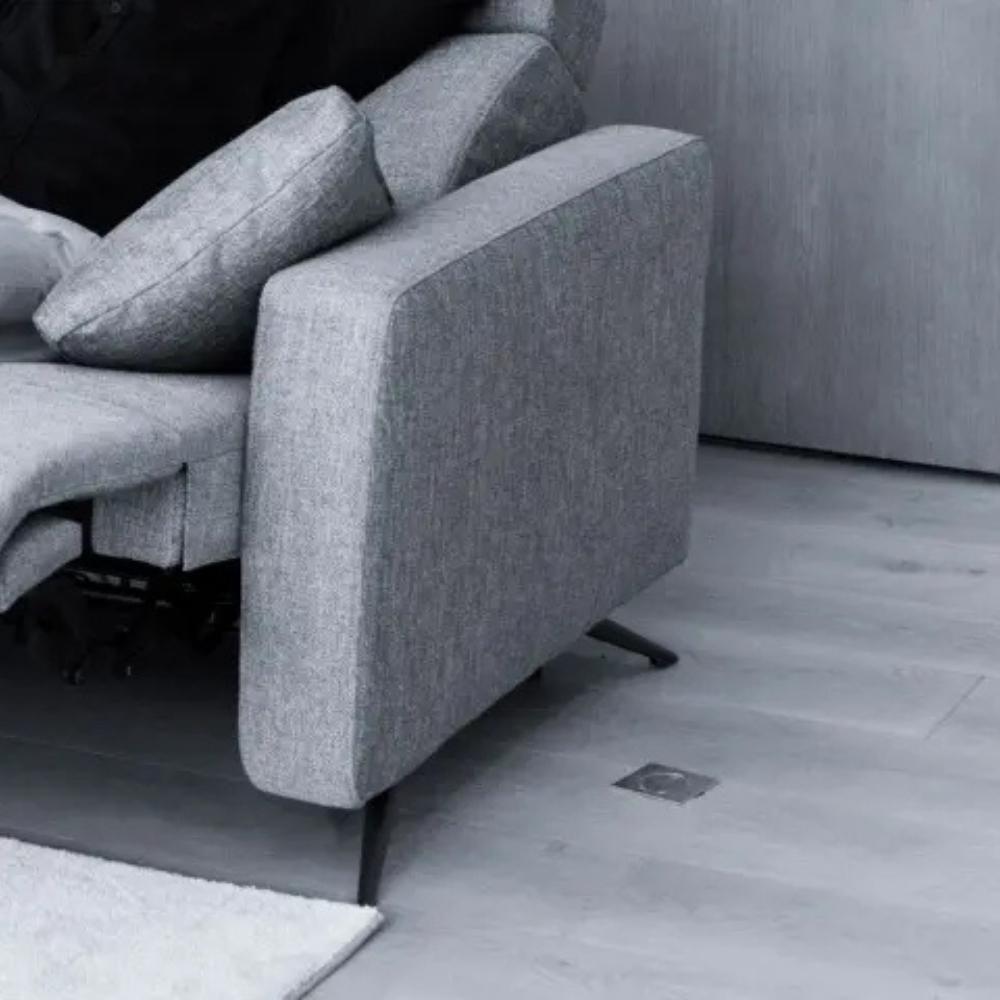 A close-up of a modern gray upholstered reclining sofa with dark metal legs, shown in a minimalist room with light gray wood flooring and a white rug.