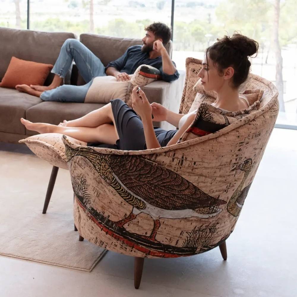 A woman relaxes in a large upholstered armchair with a bird and tree print, while a man lounges on a sofa in the background of a bright living room.