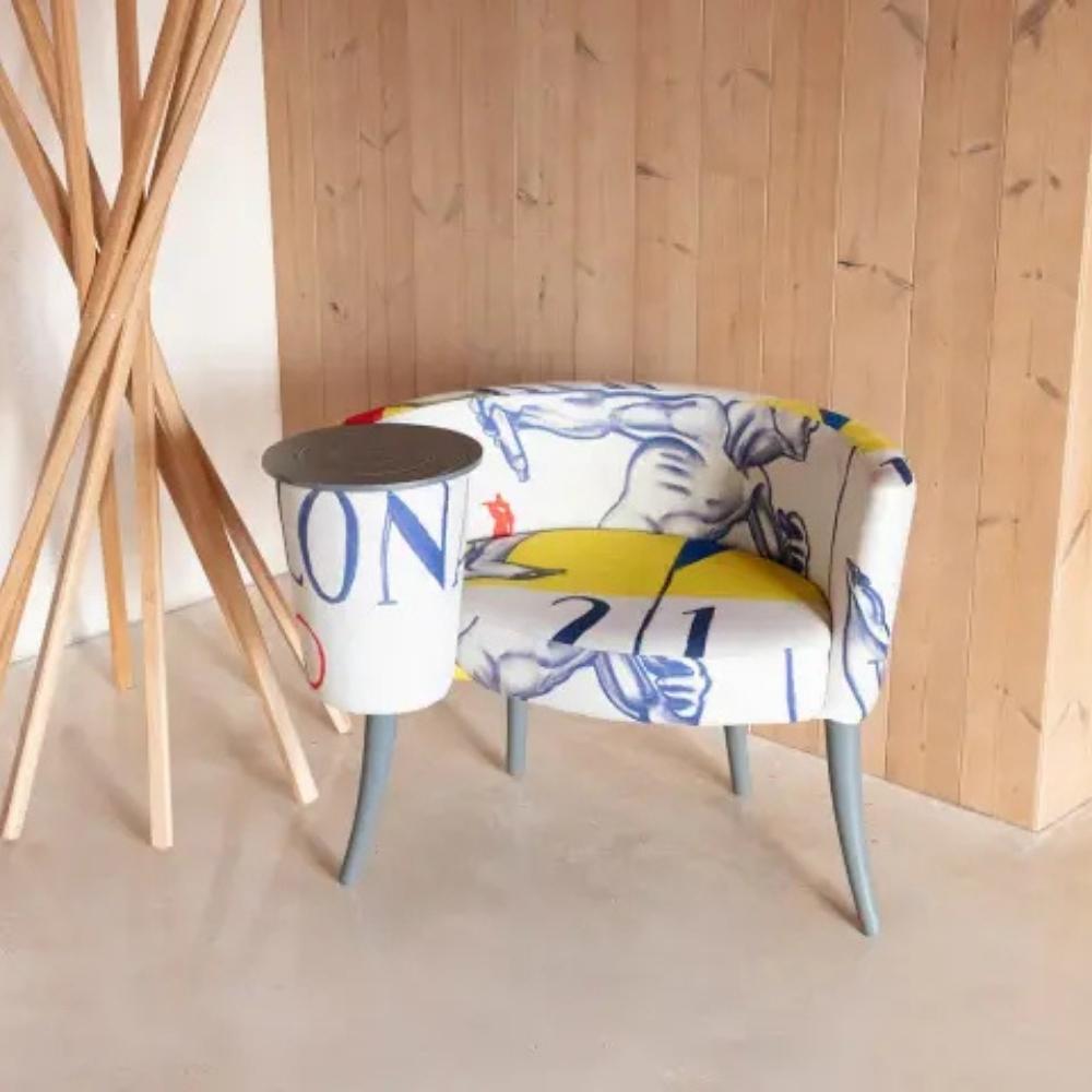 A modern upholstered accent chair with a white base and blue graphic print, featuring a cylindrical side table attached to its right arm. The chair is set against a light wood-paneled wall with a wooden ornament to one side.
