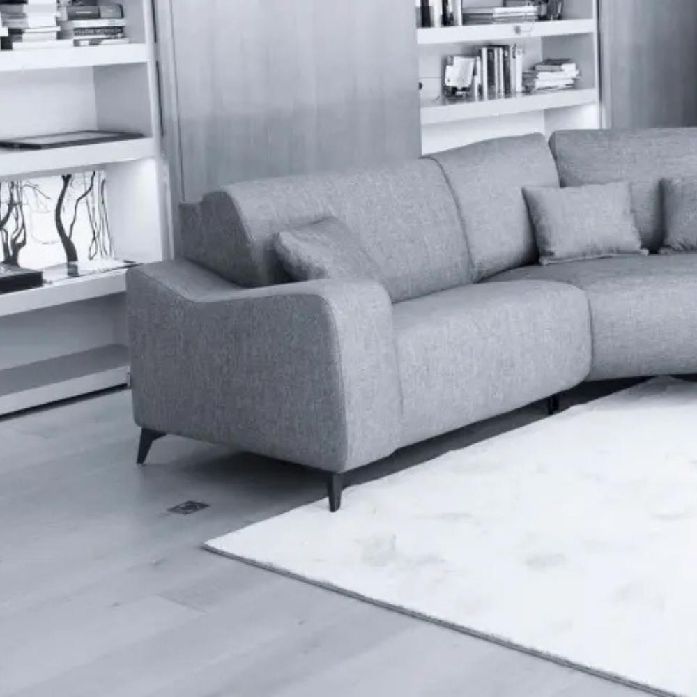 A modern gray fabric sectional sofa with dark tapered legs, styled in a minimalist living room setting with a white rug, light wood flooring, and white built-in bookshelves.