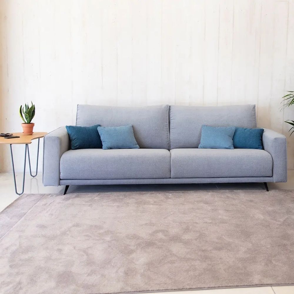A light gray fabric sofa with four throw pillows in shades of blue, styled in a minimalist living room setting with a wooden side table, potted plant, and a large textured rug.