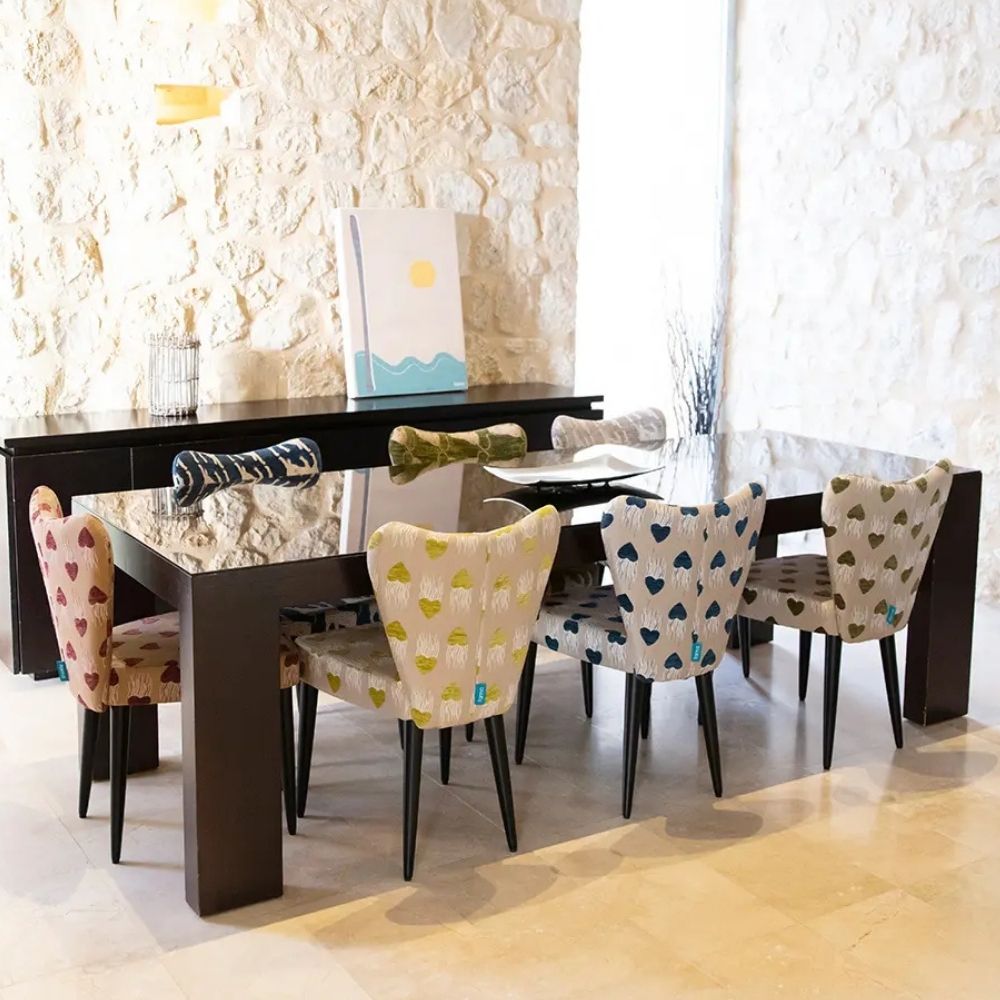 A modern dining table set with six upholstered chairs in a bright room with a stone wall. The table is dark brown with a glass top, and the chairs feature patterned fabric upholstery in cream, pink, blue, and yellow tones.