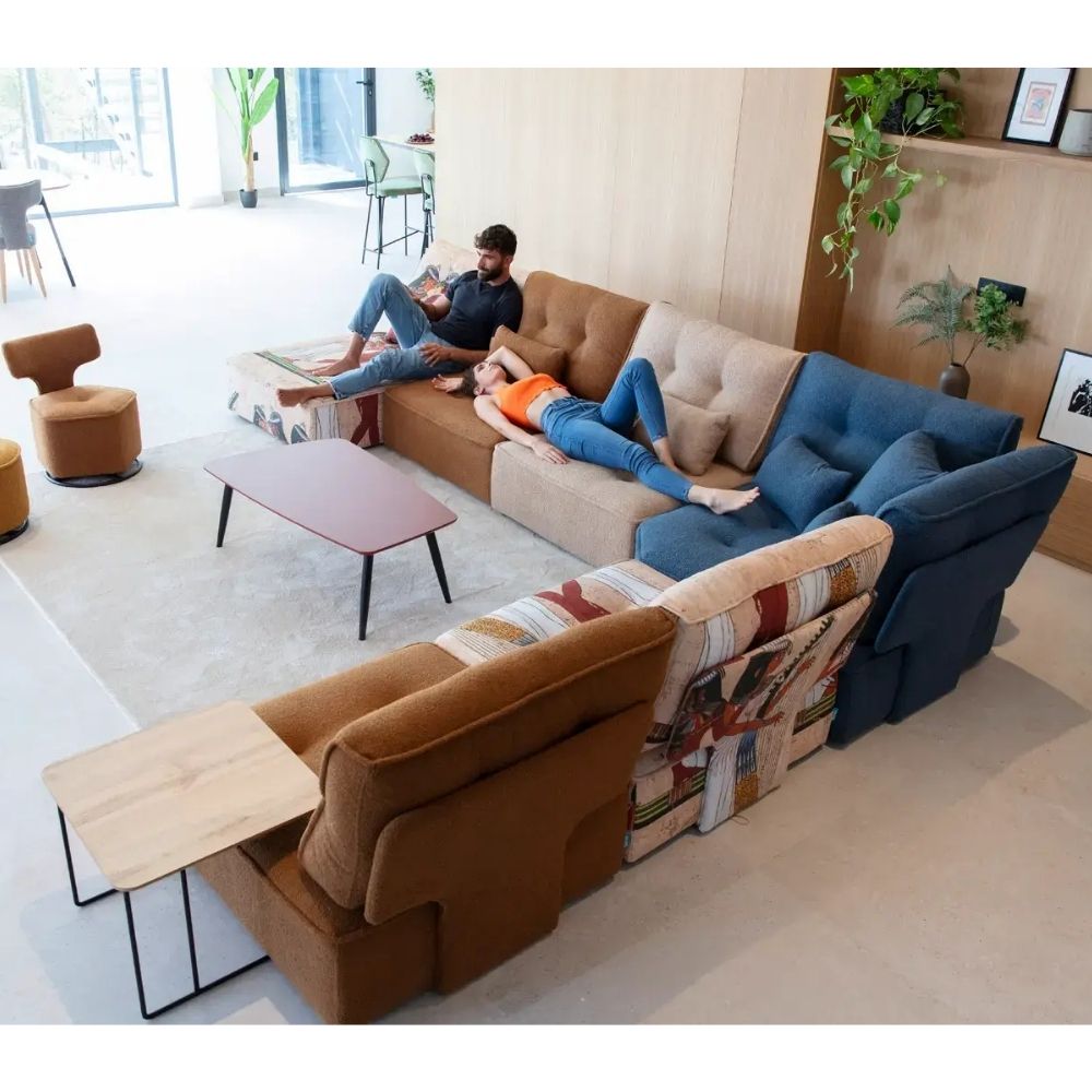 A large modular sectional sofa in a modern living room setting, featuring a brown armchair, a tan L-shaped sofa, a blue section, and a patterned accent chair. Two people are lounging on the sofa, and two small side tables are attached to the front.