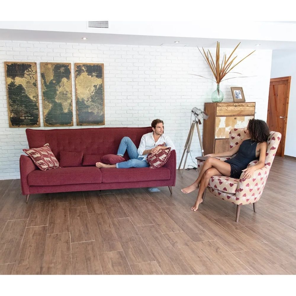 A man sits on a burgundy fabric sofa in a bright living room, while a woman relaxes in a armchair with a heart pattern and built in side table. The room features a white brick wall, wooden floors, and decorative wall art.
