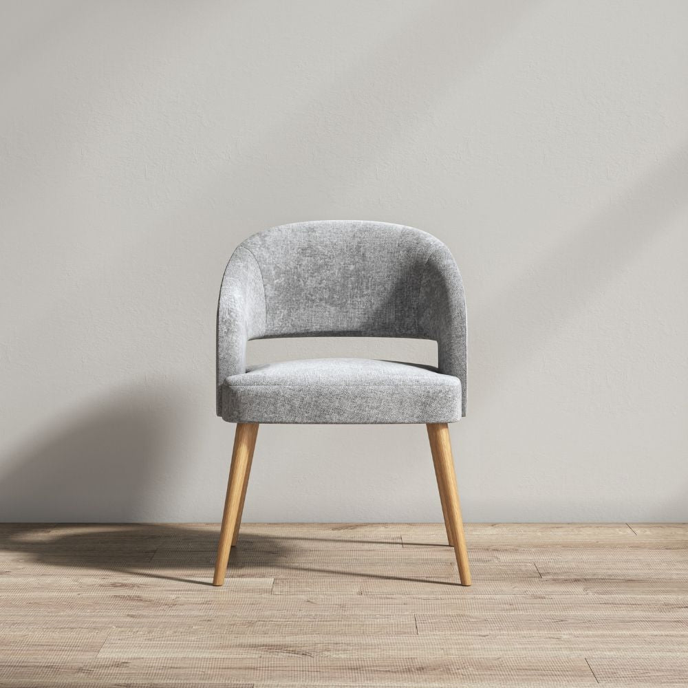 A modern grey upholstered accent chair with tapered wooden legs, set against a minimalist beige wall and light wood floor.