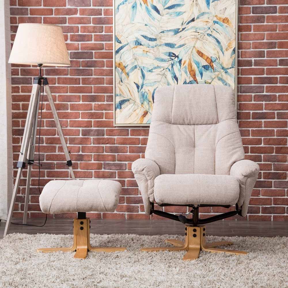 Beige armchair and footstool with a lamp and abstract painting against a brick wall.