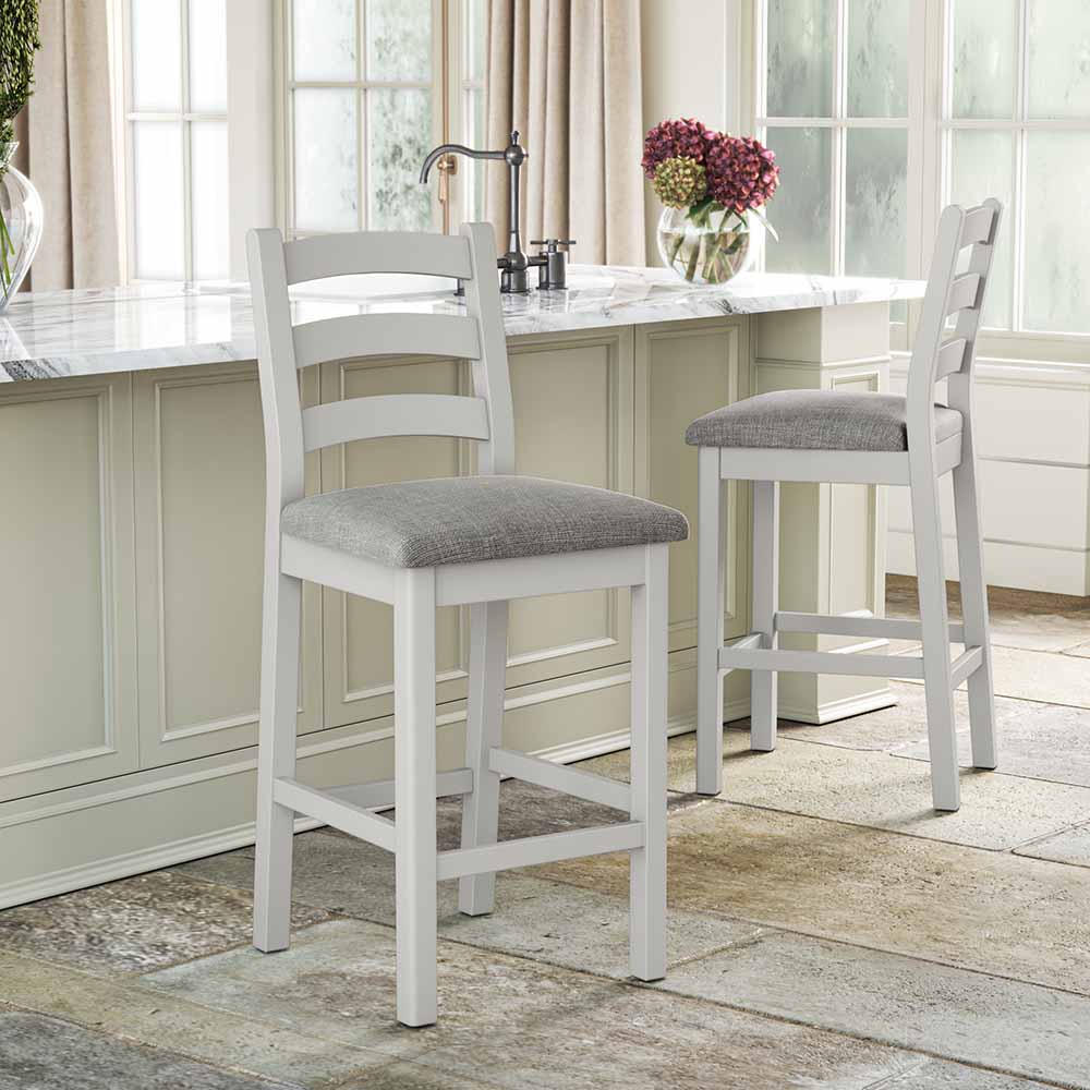 Image of two grey painted oak bar stools with grey cushions. One is facing towards a kitchen island the other is facing towards the viewer.
