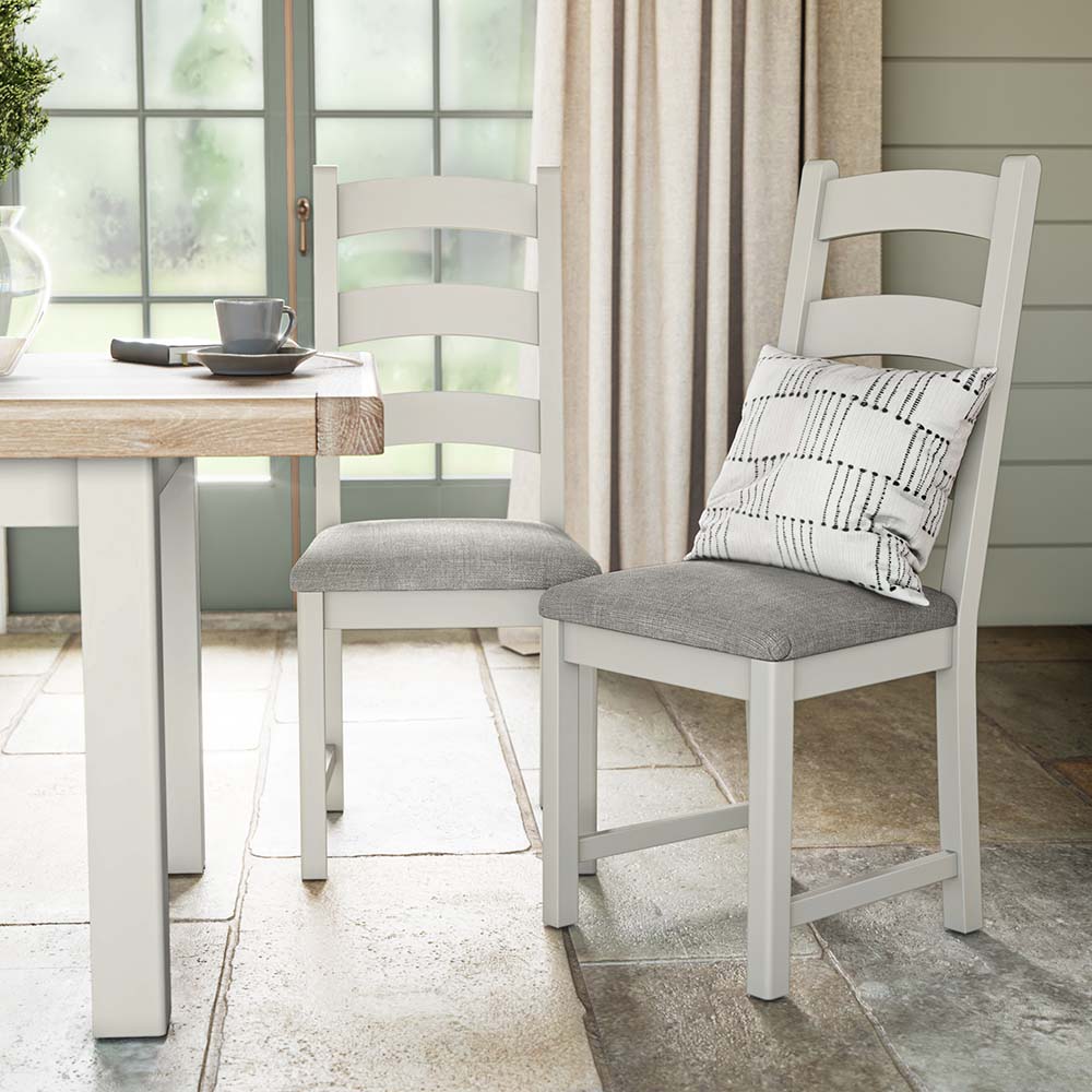 photo of two grey painted oak chairs near a matching grey painted oak dining table with grey seat cushion and a silver scatter cushion on one of the chairs. The chairs and table are set in a brightly lit litchen with big window and cream curtains
