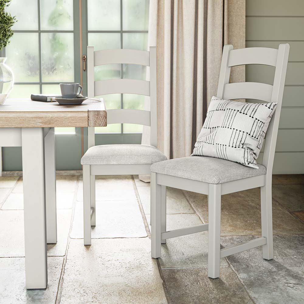 photo of two grey painted oak chairs near a matching grey painted oak dining table with beige seat cushion and a silver scatter cushion on one of the chairs. The chairs and table are set in a brightly lit kitchen with big window and cream curtains