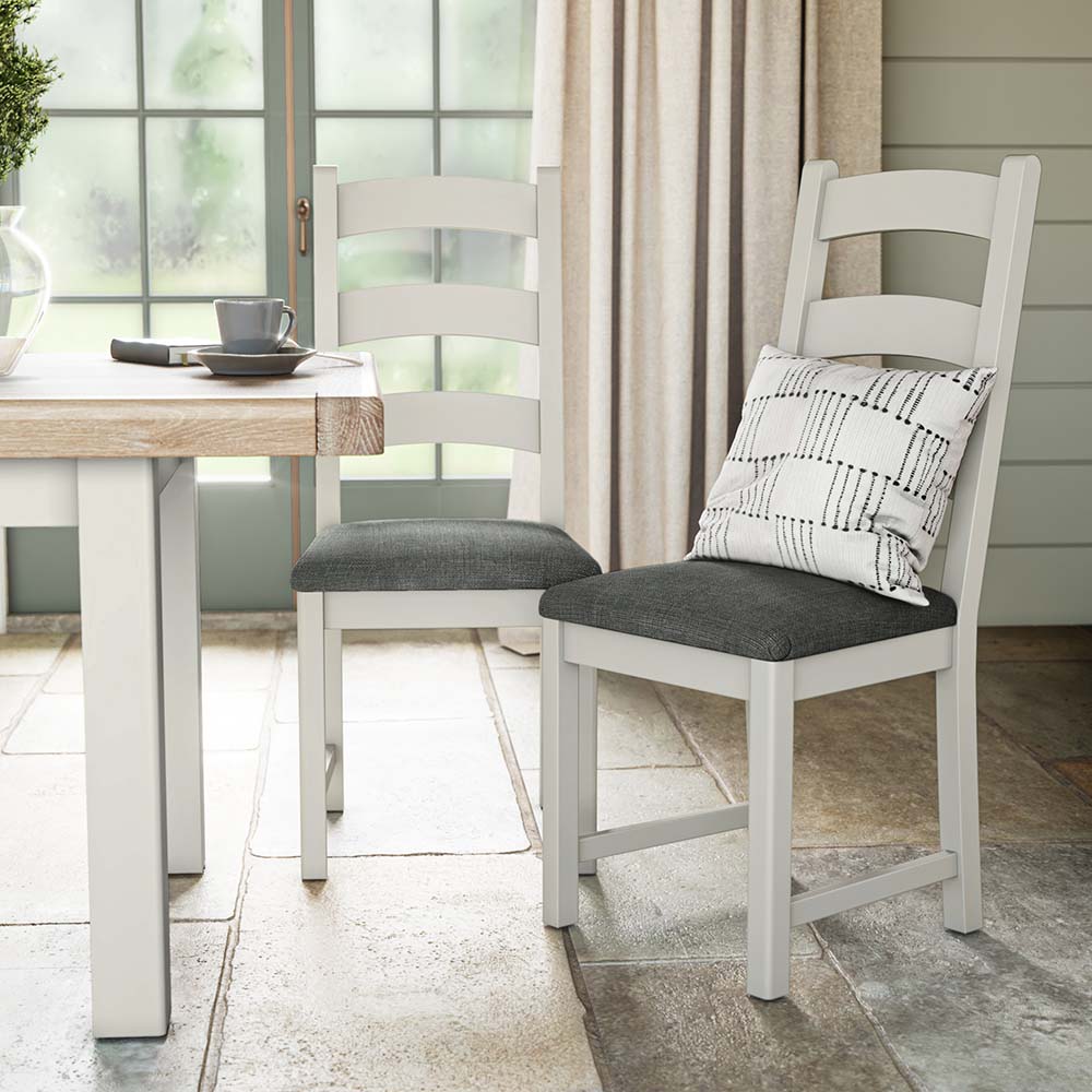 photo of two grey painted oak chairs near a matching grey painted oak dining table with charcoal grey seat cushion and a silver scatter cushion on one of the chairs. The chairs and table are set in a brightly lit kitchen with big window and cream curtains