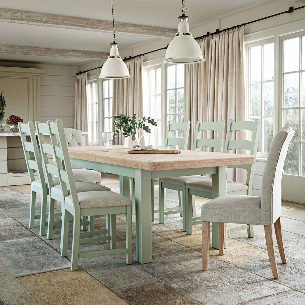 Picture of a warm neutral coloured kitchen with a sage oak extending dining table surrounded by matching sage dining chairs. 2 cream lightshades hang from the ceiling and there are plants and flowers present too.