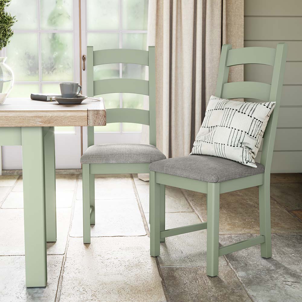 Photo of two sage painted oak chairs with grey seat cushion and a silver scatter cushion on one of the chairs. Situated near a matching sage painted oak dining table. The chairs and table are set in a brightly lit kitchen with big window and cream curtains in the background.