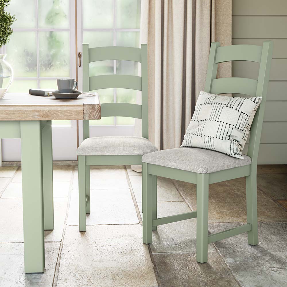 Photo of two sage painted oak chairs with beige seat cushion and a silver scatter cushion on one of the chairs. Situated near a matching sage painted oak dining table. The chairs and table are set in a brightly lit kitchen with big window and cream curtains in the background.